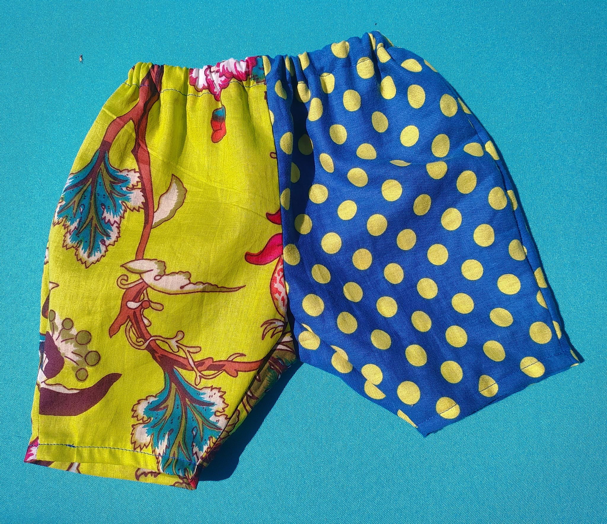 A tiny pair of baby pants laid flat on a turquoise background — one leg in chartreuse floral fabric, the other in blue with yellow polka dots