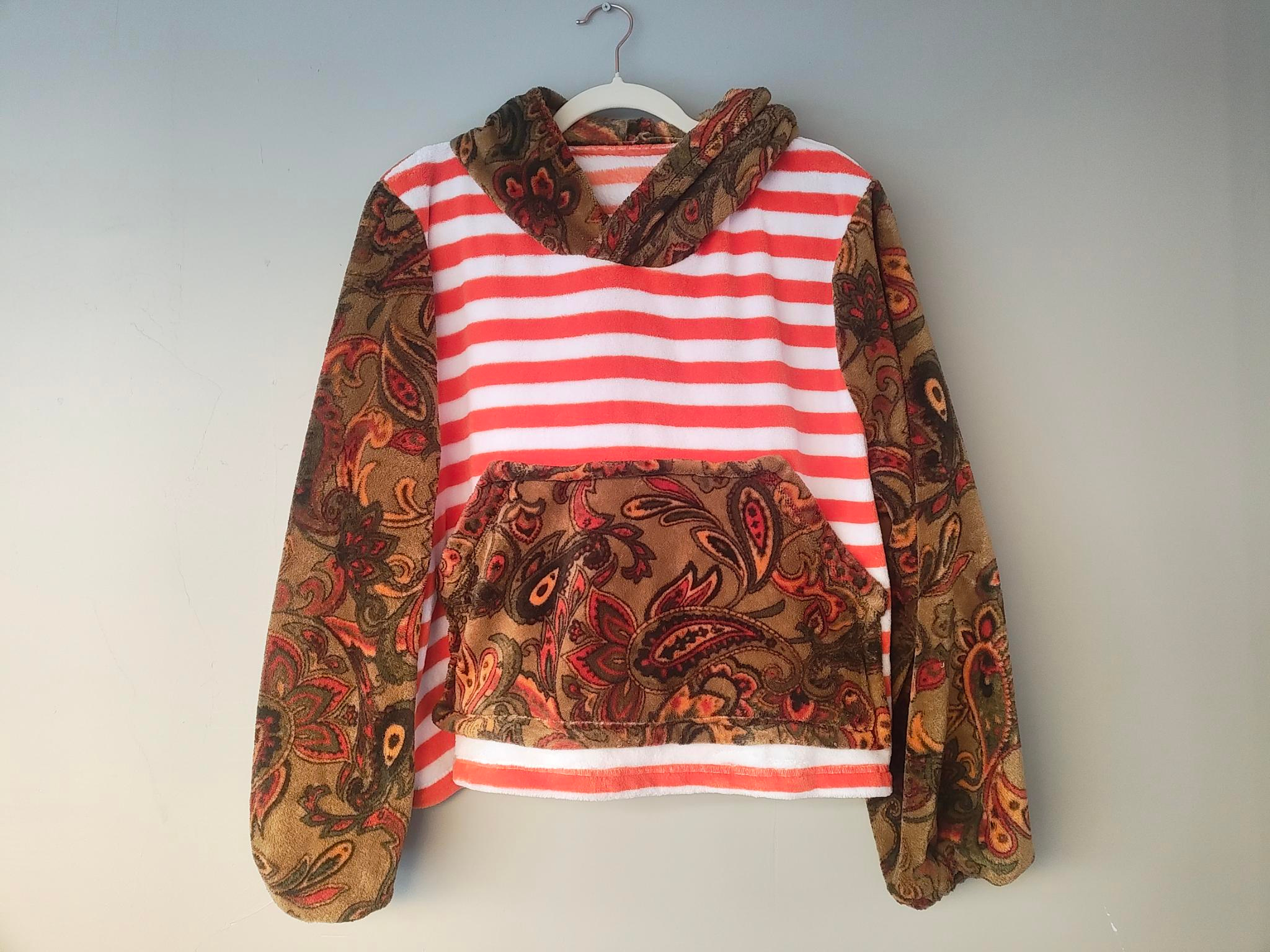 A fleece hoodie on a wire hanger against a white wall — coral-and-white striped body, with sleeves, hood, and kangaroo pocket in a brown paisley print